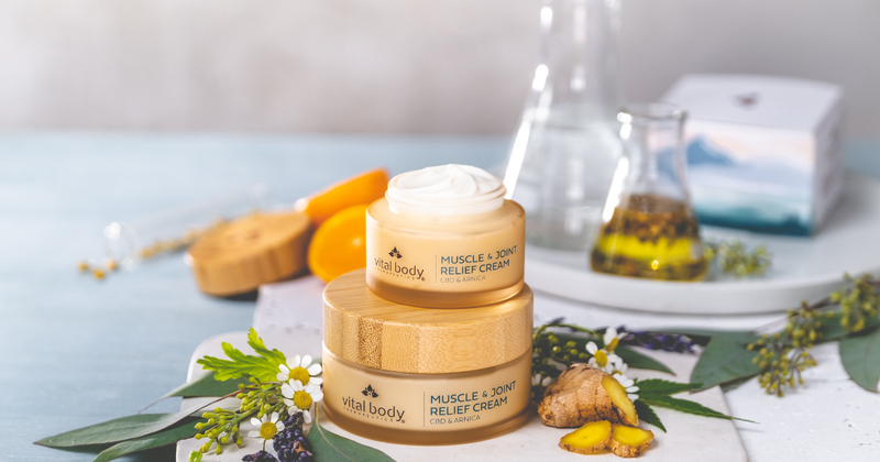 Vital Body Therapeutics' Muscle & Joint Relief Cream in two stacked jars with bamboo lids, the top jar open, surrounded by chamomile, lavender, ginger and eucalyptus, with glass flasks of infusing oil behind.