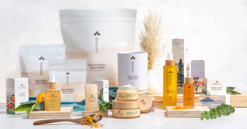 Vital Body Therapeutics' product line on a light surface, with bath soak pouches, boxed cartons, gold oil bottles, cream jars with bamboo lids, dried flowers and eucalyptus.
